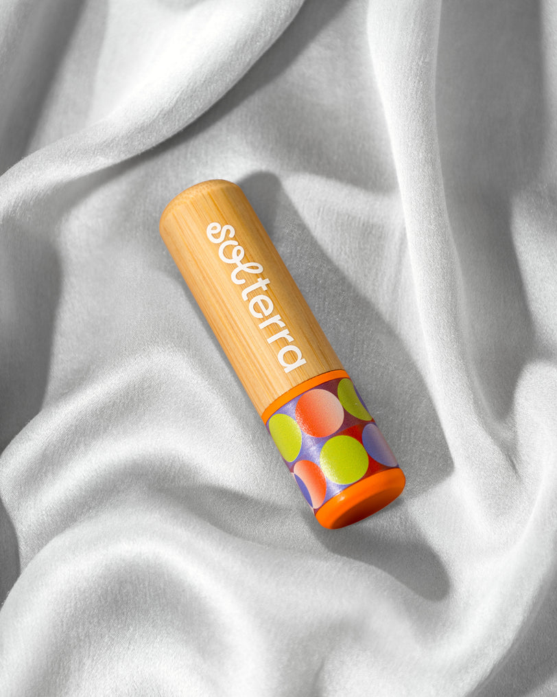 A tube of soterra lip balm with a light wood-grain body, white brand lettering, and a colorful polka-dot patterned cap, resting on draped white fabric.