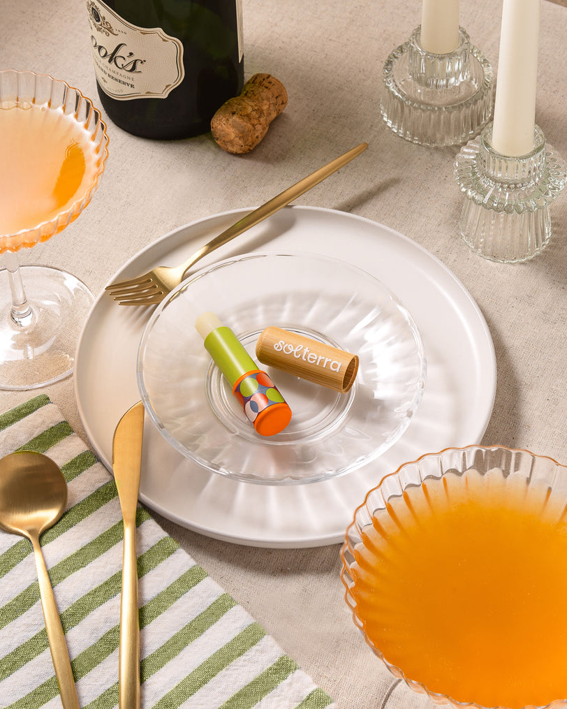A styled tabletop scene with a small clear glass dish holding a green and orange striped lip balm tube labeled 'softerra' and a tan cylindrical item. The setting includes white plates, gold-toned cutlery, candlesticks, glasses of orange drink, a striped napkin, and a wine bottle in the background.
