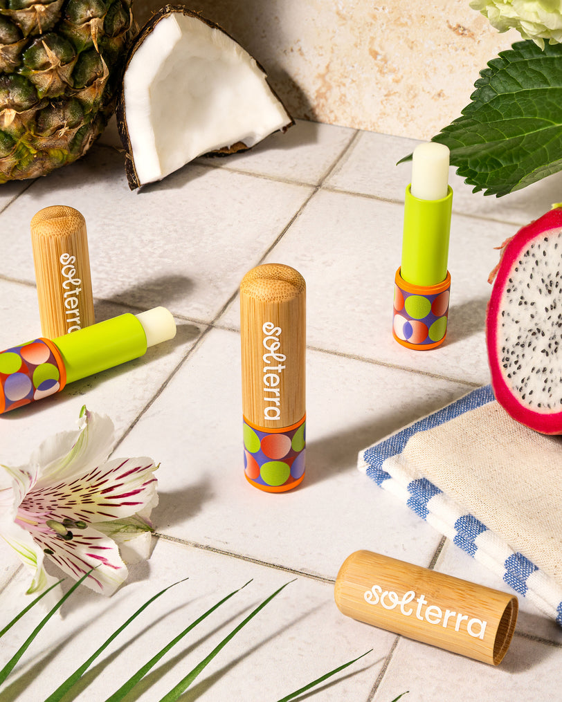 Solterra lip balm tubes on a light wooden surface with tropical fruits and leaves.