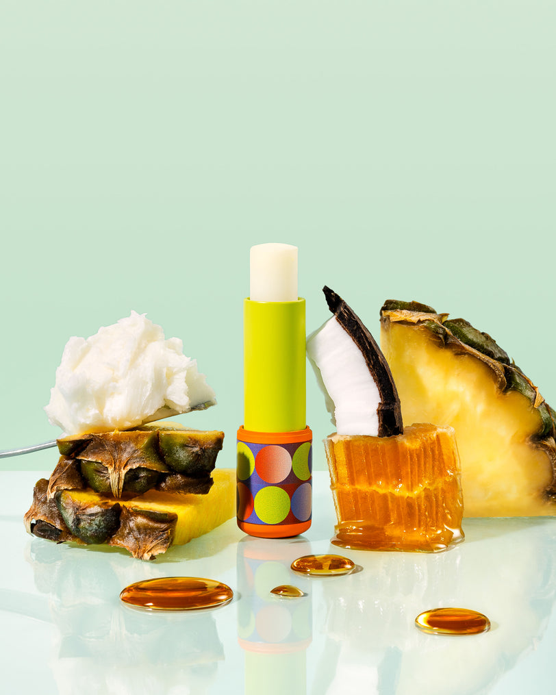 Lip balm with colorful design, pineapple slices, and a glass container on a light green background